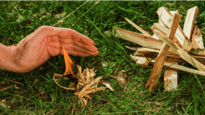 a person shielding lit kindling from the wind next to a small pile of wood ready to be lit.
