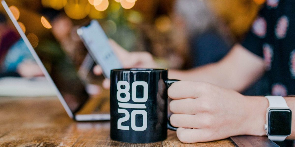 a person working on a laptop holding a mug with "80/20" printed on it