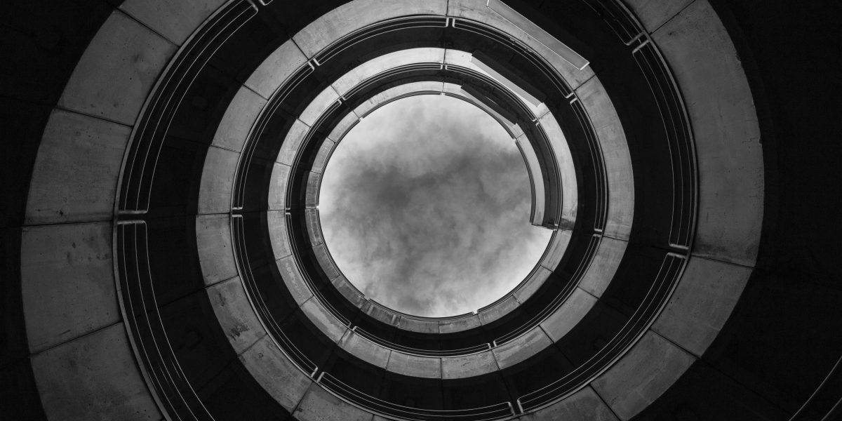 a black and white photo looking up a spiraling parking ramp toward a cloudy sky